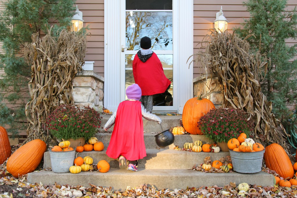 Two young children dressed up in costumes are waiting at a house for candy while Trick-or-Treating on Halloween.